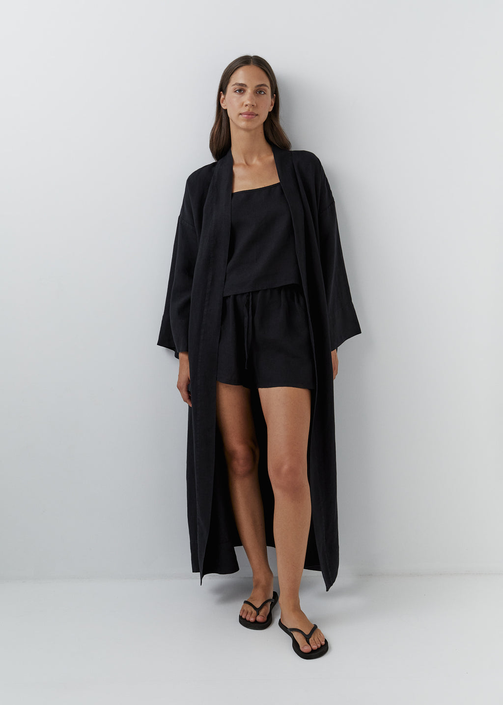 A woman stands against a light gray studio wall wearing a long black linen kimono robe with a waist belt and wide sleeves, paired with black flip-flop sandals.
