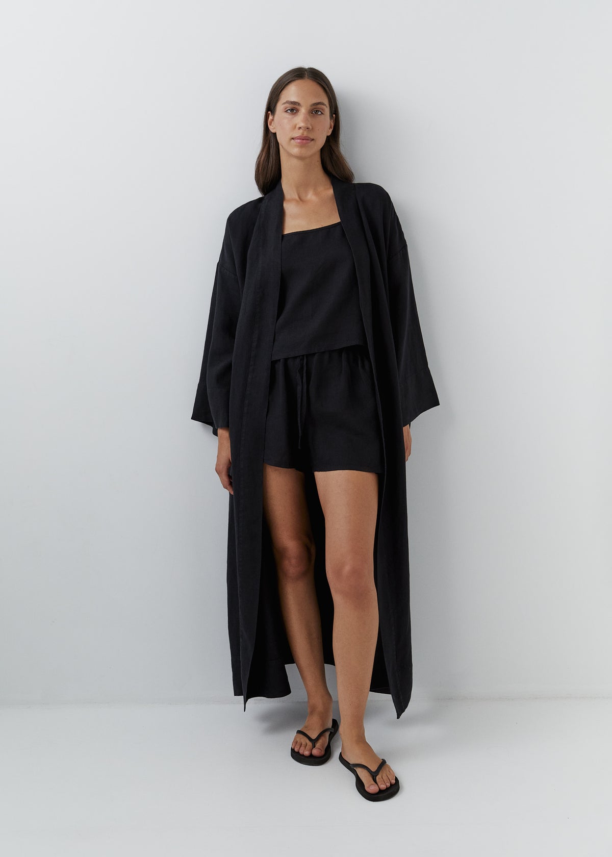 A woman stands against a light gray studio wall wearing a long black linen kimono robe with a waist belt and wide sleeves, paired with black flip-flop sandals.