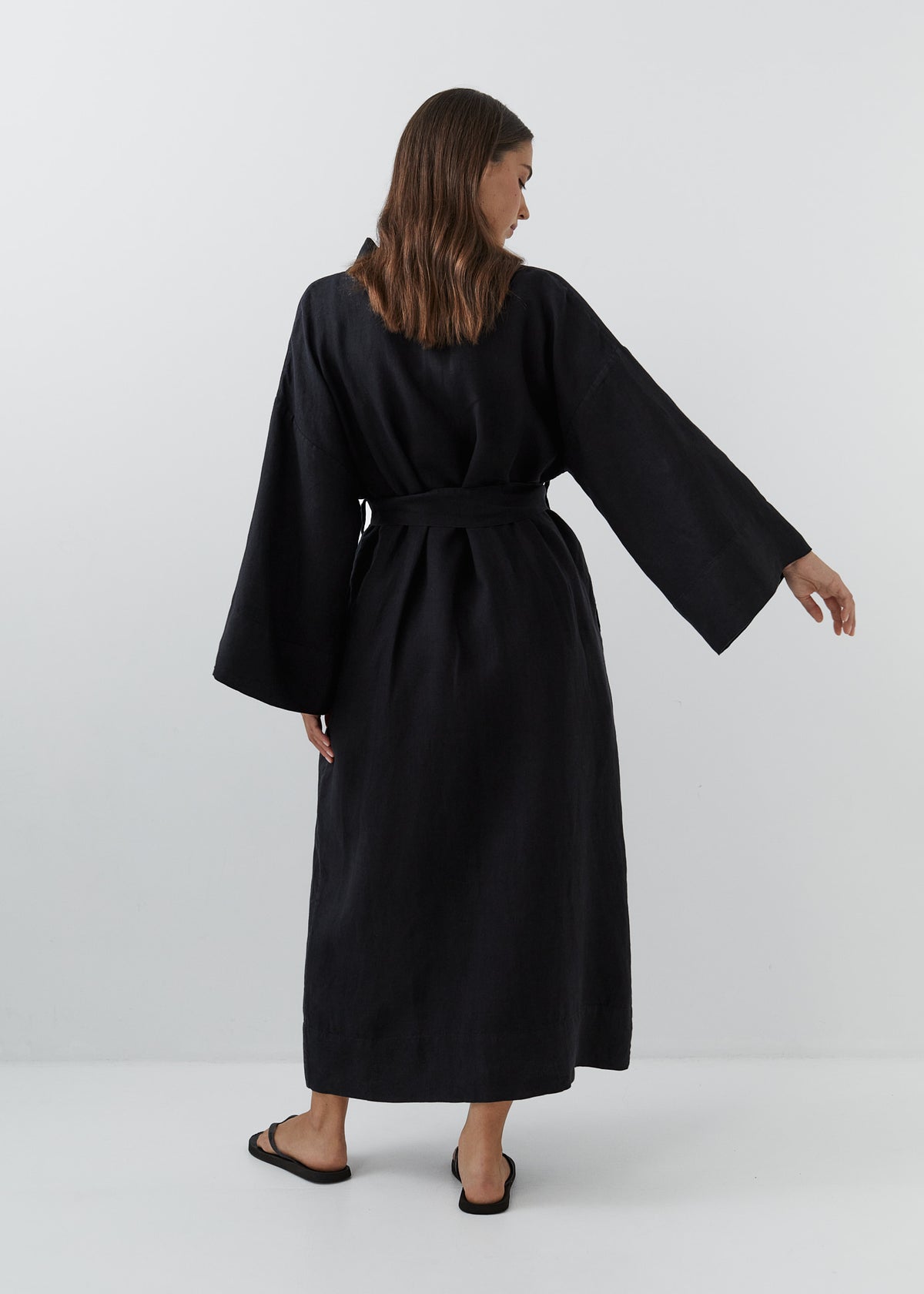 A woman wearing a long black linen robe with wide sleeves and a matching waist tie, shown from the back against a plain light gray studio background.