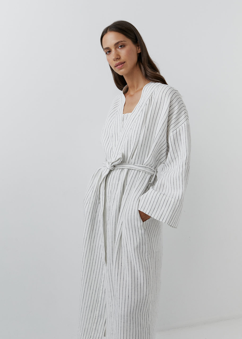 A woman wearing a white robe with thin vertical black stripes, standing against a plain light gray studio background.