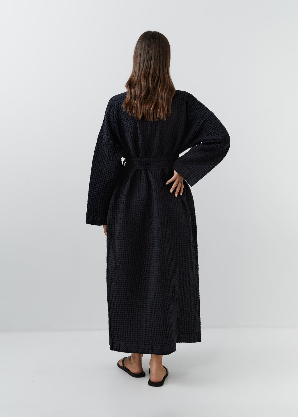 A woman viewed from behind wearing a long black waffle linen robe with a tie belt, standing against a plain light gray studio background.