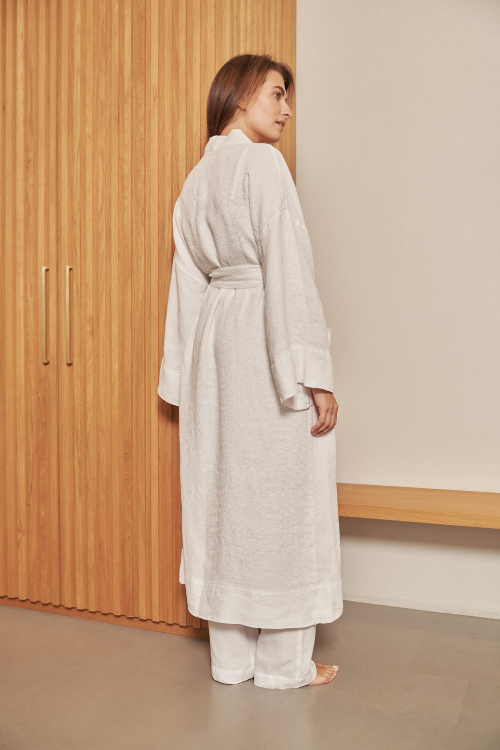 A woman modeling a long white waffle linen robe with wide sleeves and a tie belt, shown from the back and side.