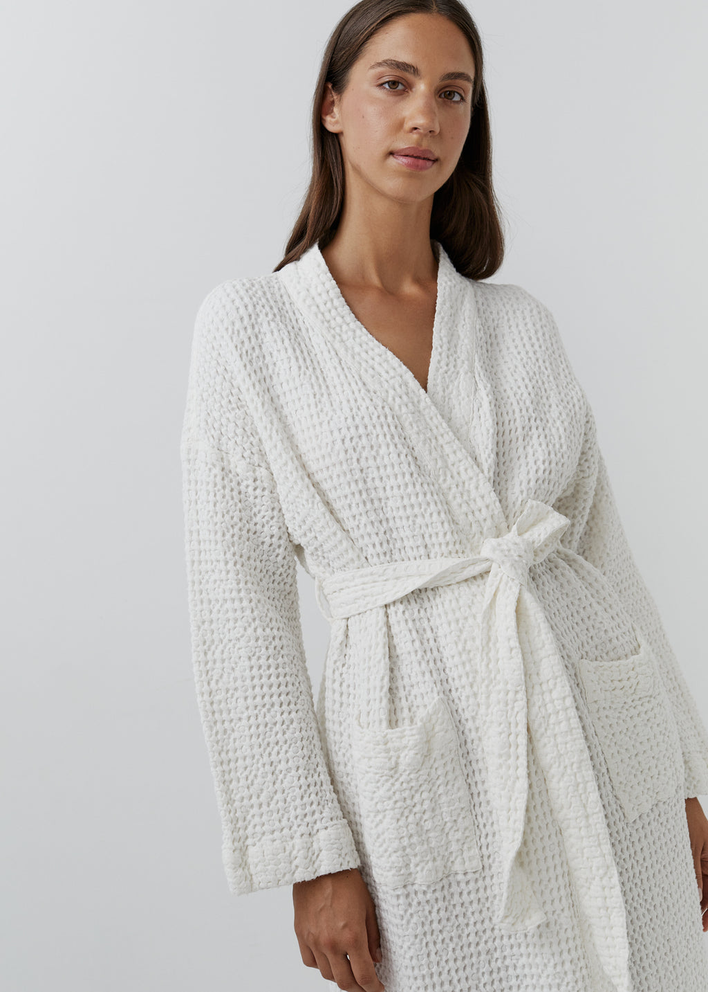 A woman wearing a white waffle-textured robe with a tie waist belt, photographed against a plain light gray studio background.