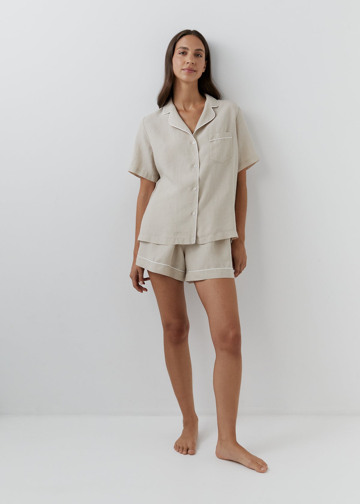 A woman modeling a light beige linen pajama set consisting of a short-sleeve button-up shirt and matching shorts, standing against a plain white studio background.