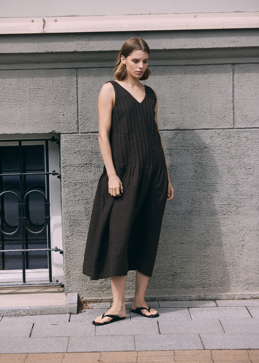 Full-length view of a woman in a dark brown sleeveless linen dress with a V-neckline, textured pleated front and loose A-line silhouette, paired with black flat sandals.