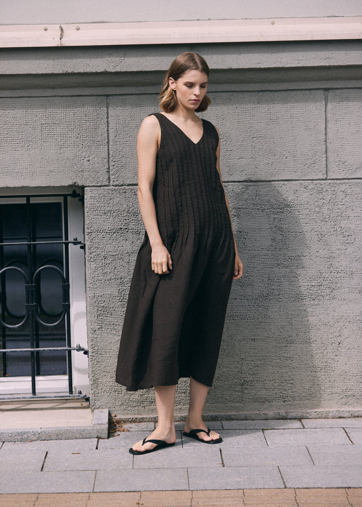 Full-length view of a woman in a dark brown sleeveless linen dress with a V-neckline, textured pleated front and loose A-line silhouette, paired with black flat sandals.