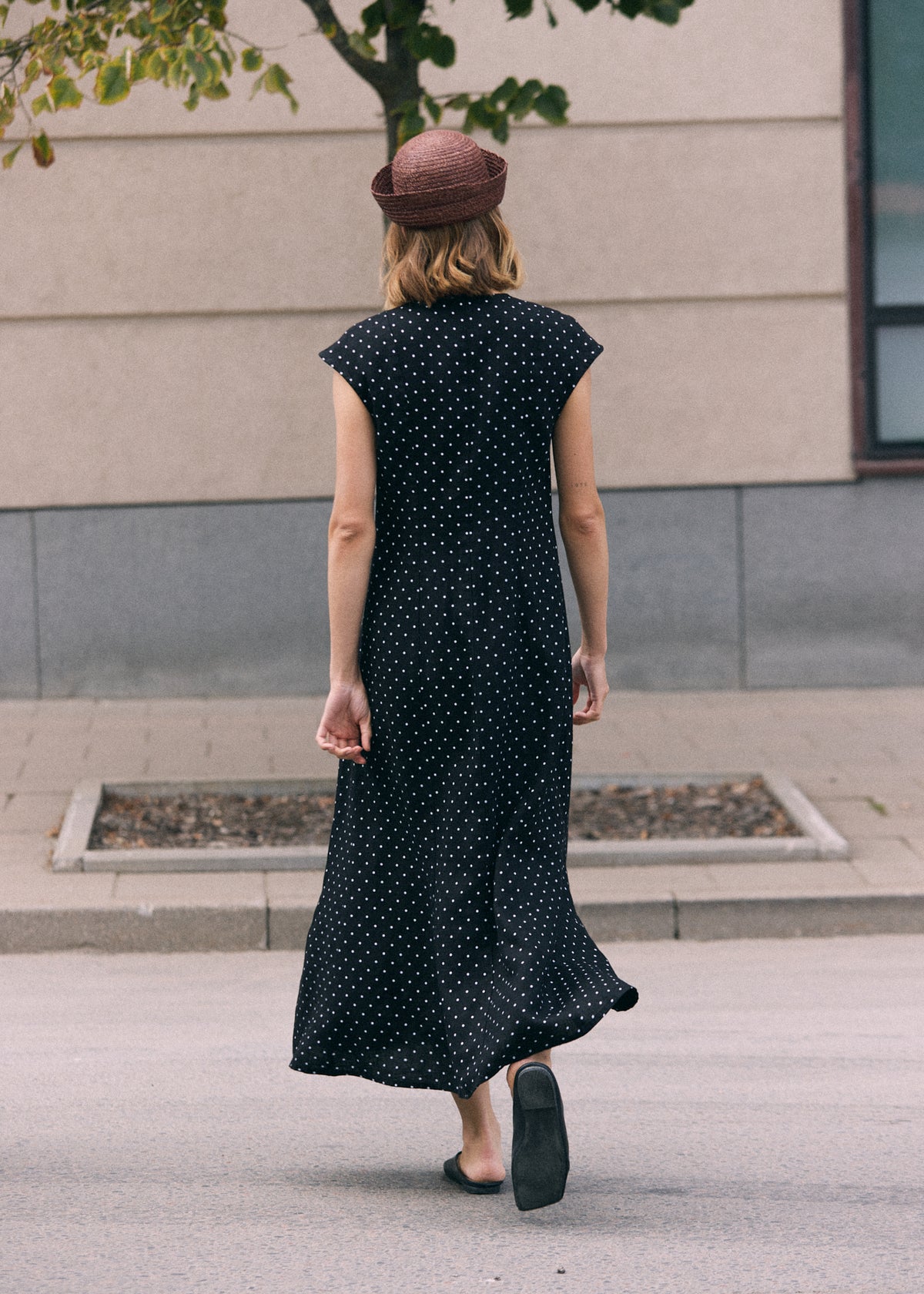 Woman walking outdoors in a black linen maxi dress with white polka dots and short cap sleeves, shown from the back and styled with a brown hat and black shoes.