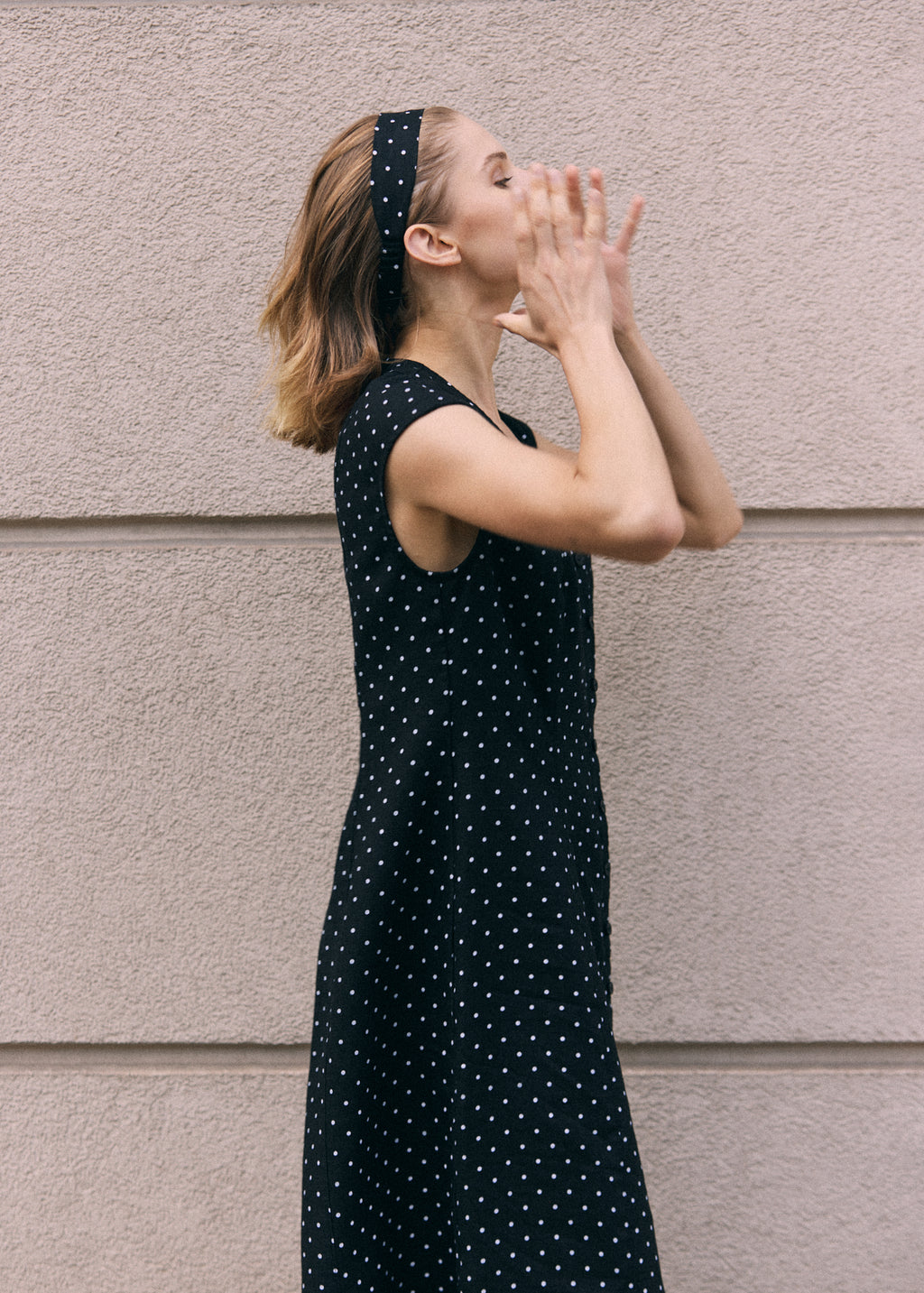 Woman wearing a black linen maxi dress with white polka dots and a matching polka dot headband, shown from the side against a neutral textured wall.