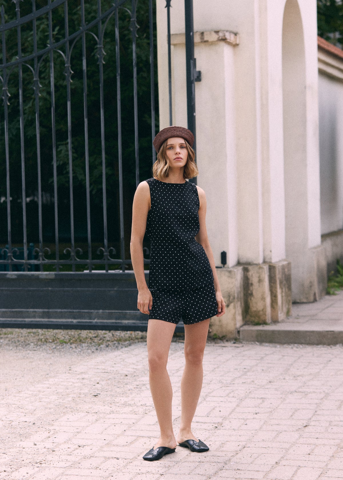 A woman stands outdoors in front of a cream-colored building and black metal gate, wearing a black sleeveless top with white polka dots and matching shorts, paired with a brown beret and black flat shoes.
