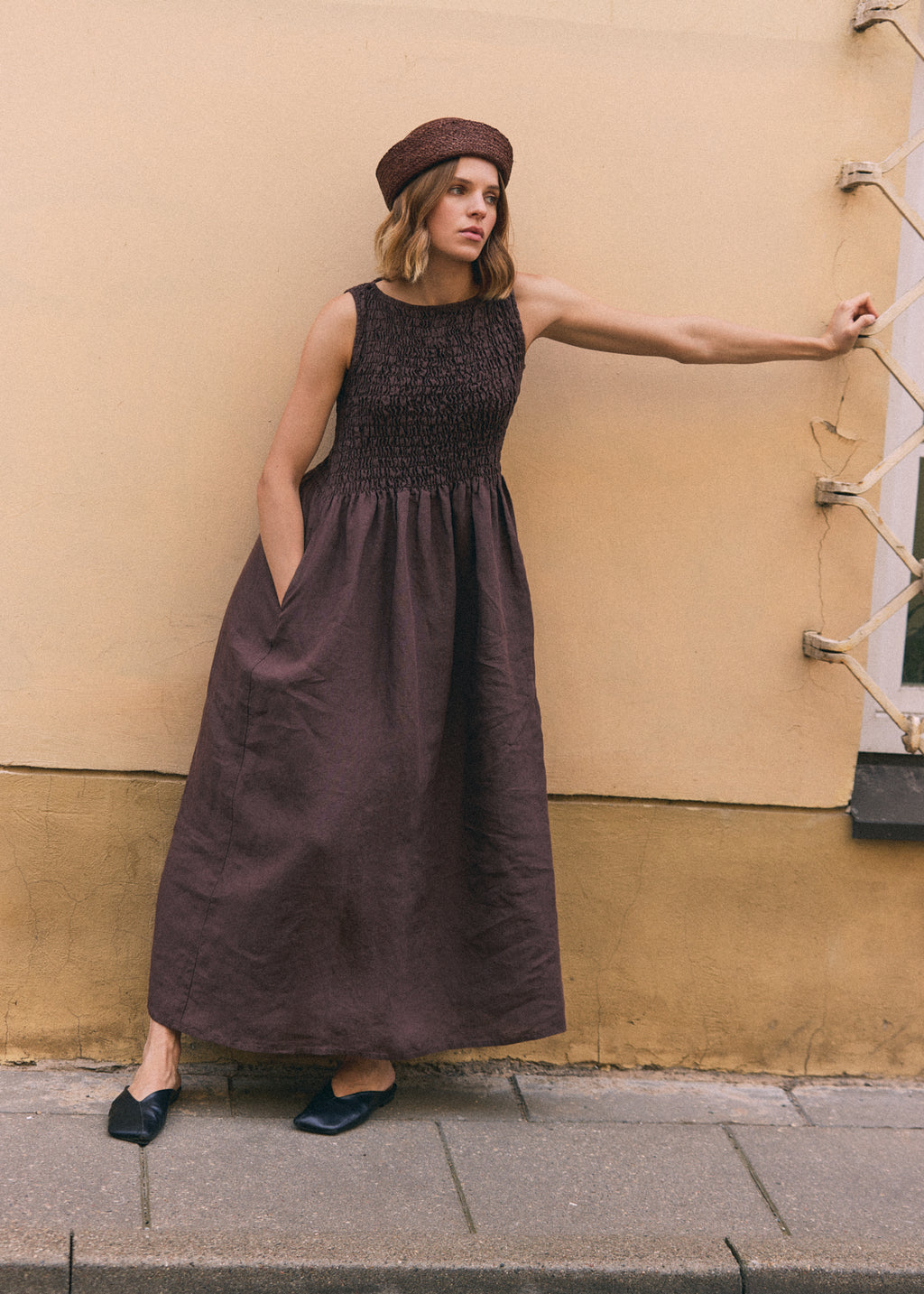 Woman in a dark brown sleeveless linen maxi dress with a fitted shirred bodice, full flowing skirt and practical side pockets, photographed against a warm neutral wall.