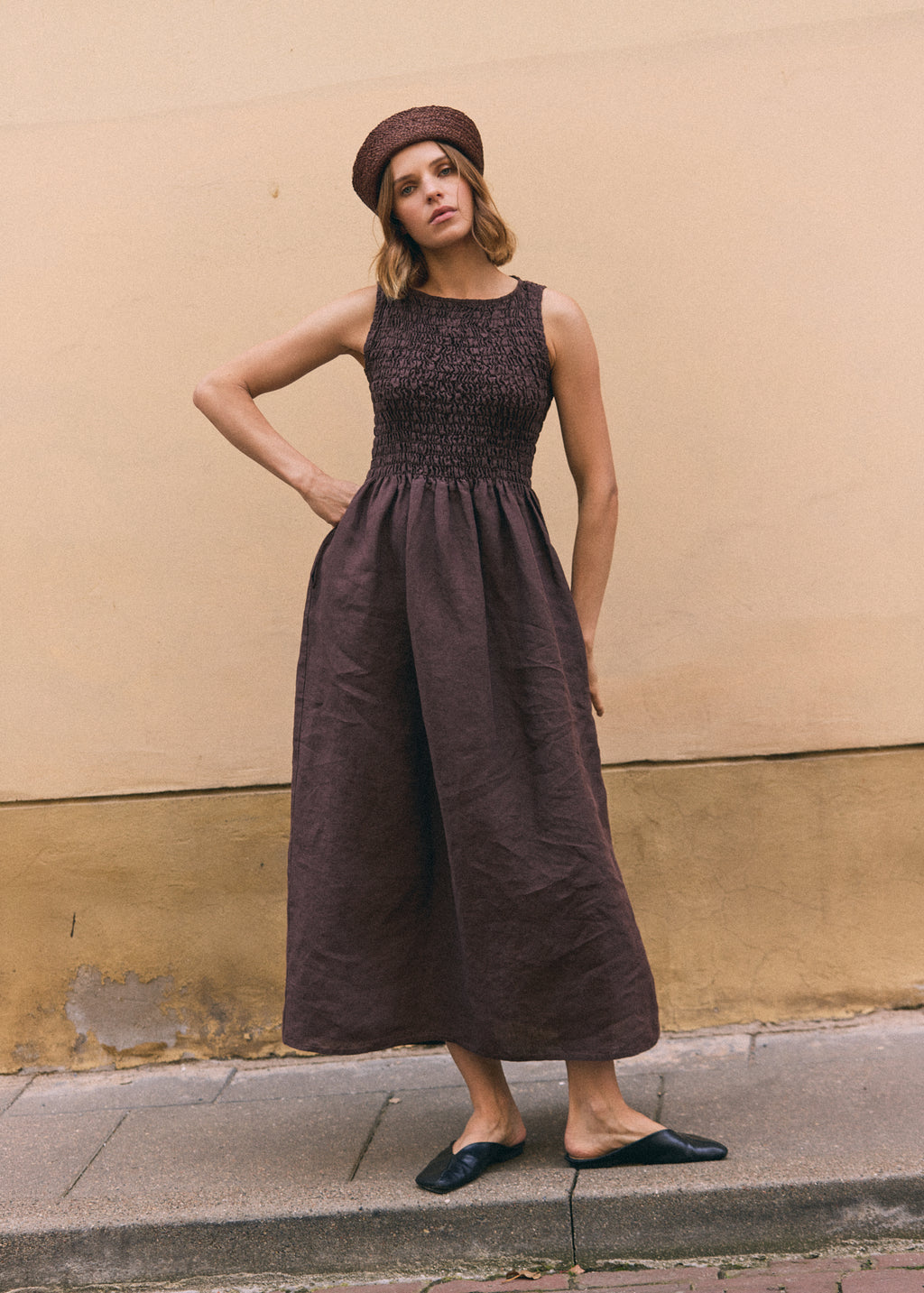 Woman wearing a dark brown linen maxi dress with a shirred bodice, sleeveless design and flowing gathered skirt, styled with a matching brown woven hat and black flats.