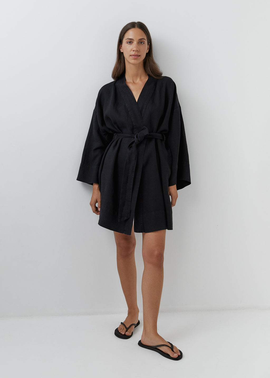 A woman modeling a short black linen kimono robe with wide long sleeves, side pockets and a tie belt.