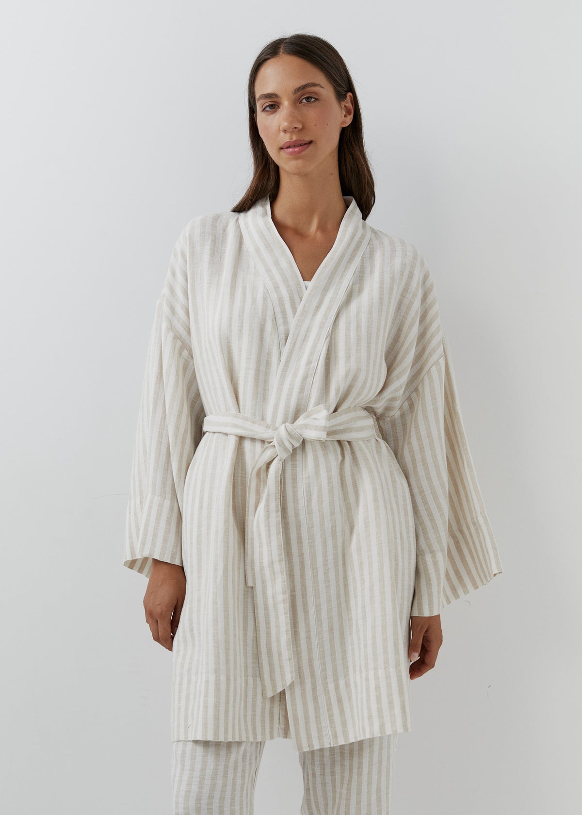 A woman modeling a cream and white vertically striped linen robe with wide long sleeves and a tie belt, paired with matching striped trousers against a plain light gray background.