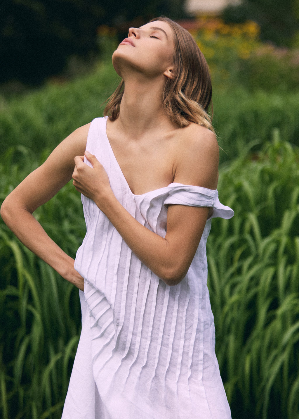 Close-up of a woman in a white linen maxi dress with a V-neckline and vertical pleated detailing, posing against a lush green garden background.