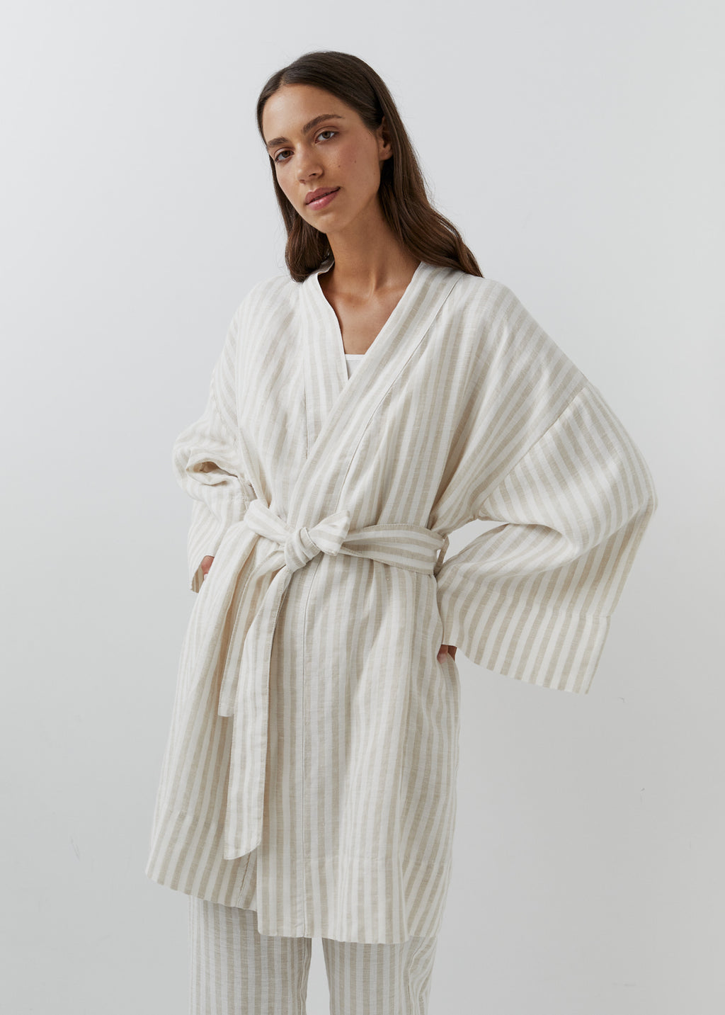 A woman modeling a cream and white vertically striped linen robe with wide long sleeves, side pockets, and a tie belt.