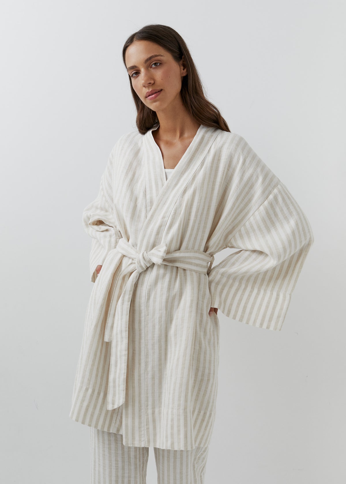 A woman modeling a cream and white vertically striped linen robe with wide long sleeves, side pockets, and a tie belt.