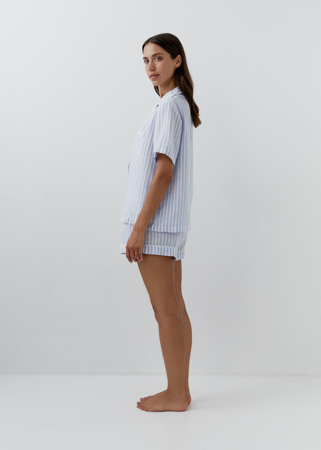 A woman stands in profile wearing a light blue and white vertically striped short-sleeve pajama shirt and matching shorts against a plain light gray studio background.