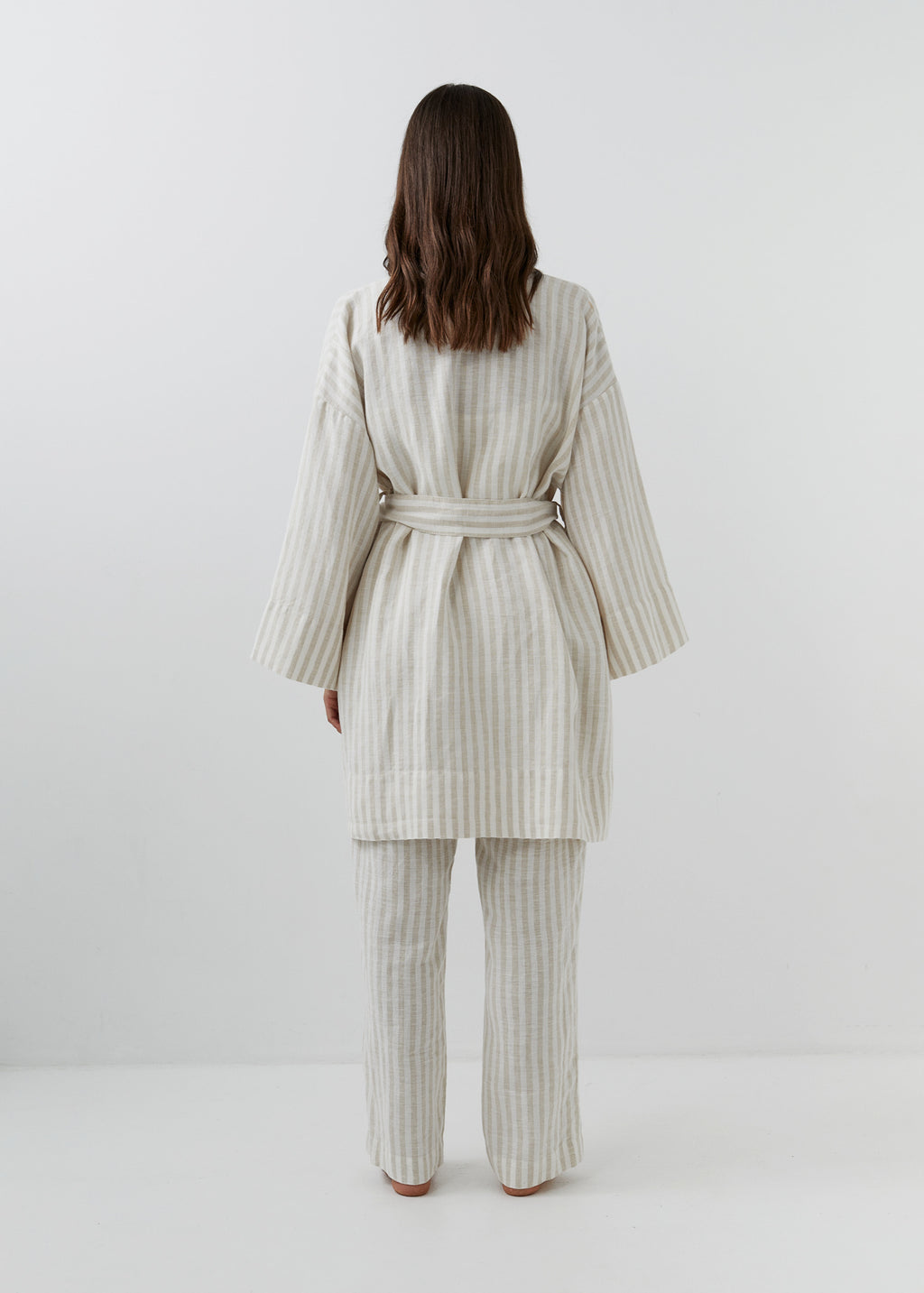 A woman modeling a cream and white vertically striped linen robe with wide long sleeves and a tie belt, shown from the back with matching striped trousers.
