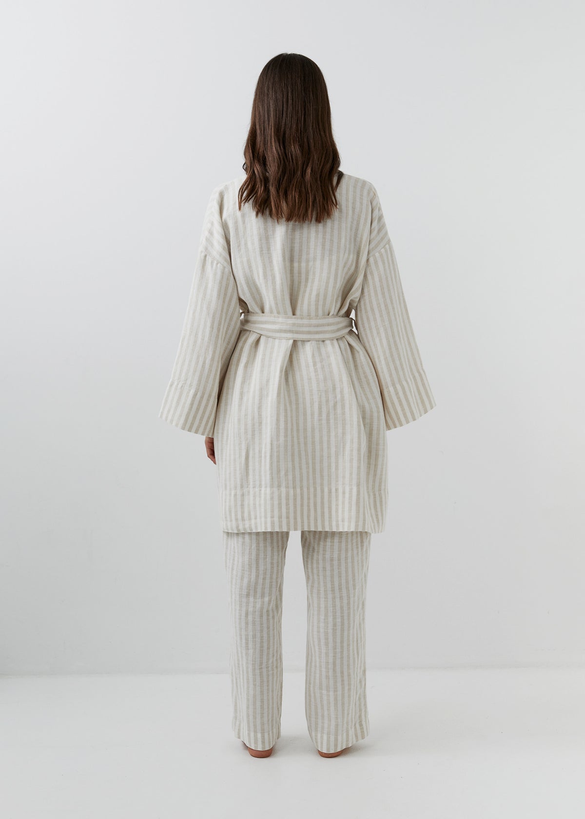 A woman modeling a cream and white vertically striped linen robe with wide long sleeves and a tie belt, shown from the back with matching striped trousers.