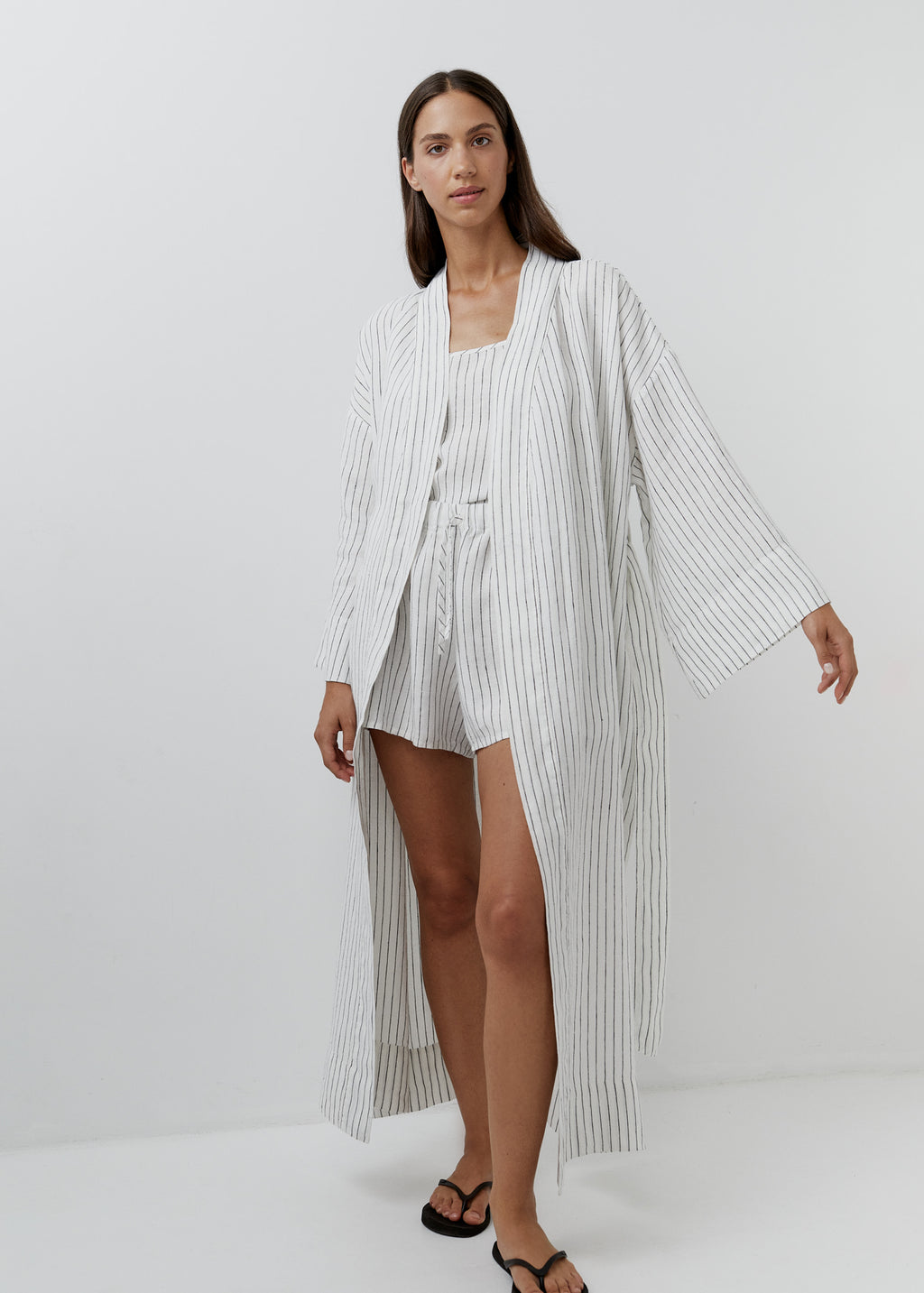 A woman modeling a white linen robe and black vertically striped lounge set consisting of a long open-front robe and matching shorts, paired with black sandals, against a plain light gray studio background.