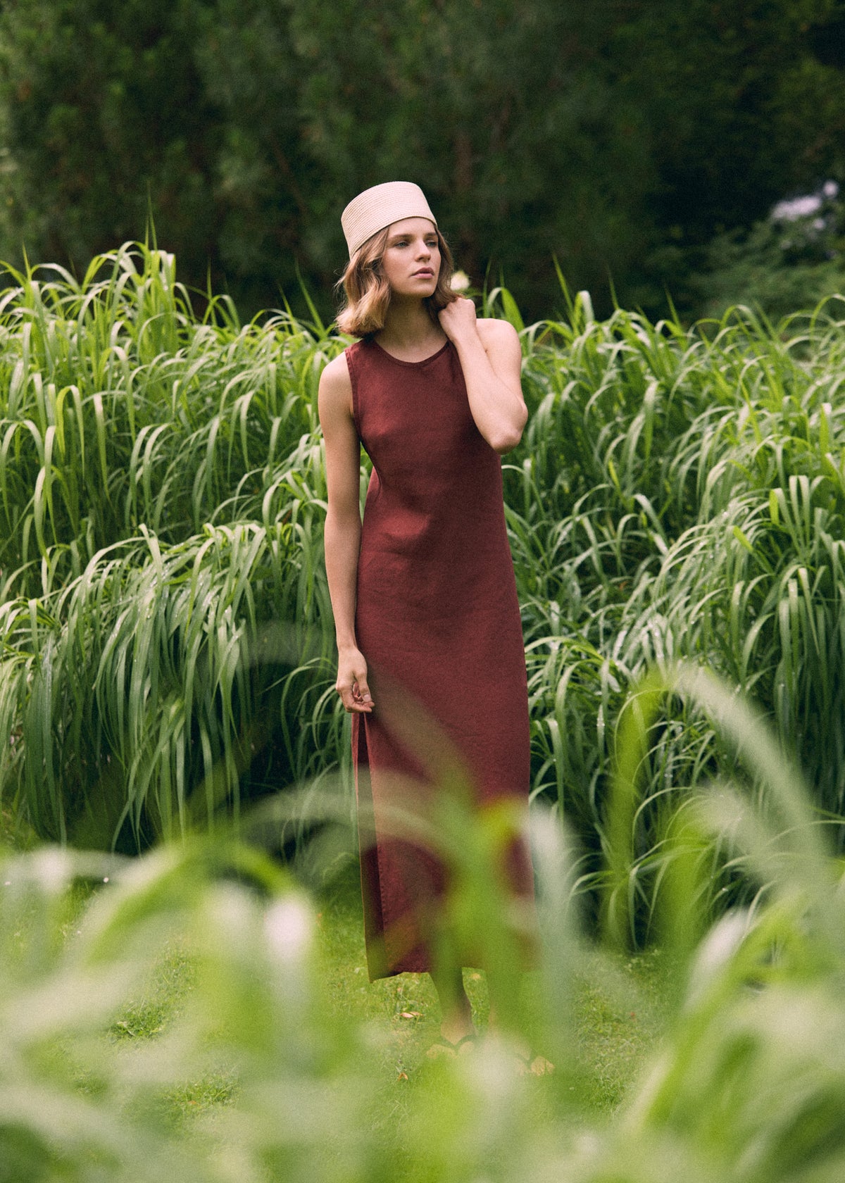 Woman in a long terracotta linen dress and light woven hat, standing among tall ornamental grasses with soft greenery in the foreground.