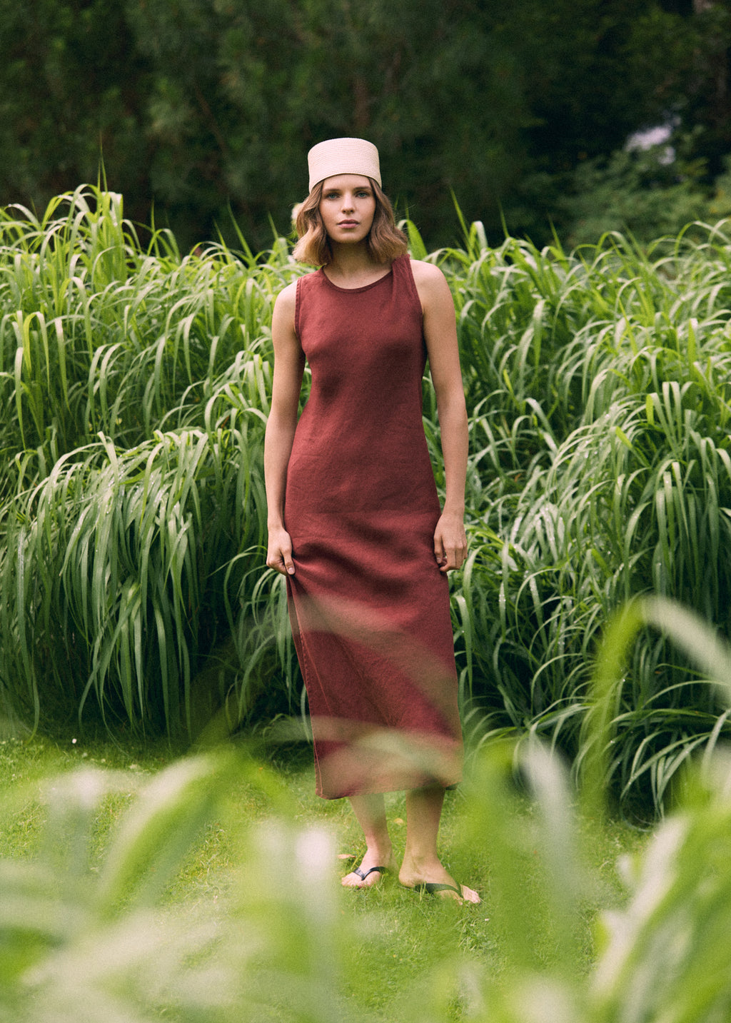 Woman wearing a terracotta sleeveless linen maxi dress and a light woven hat, standing among tall green grasses in a lush garden.