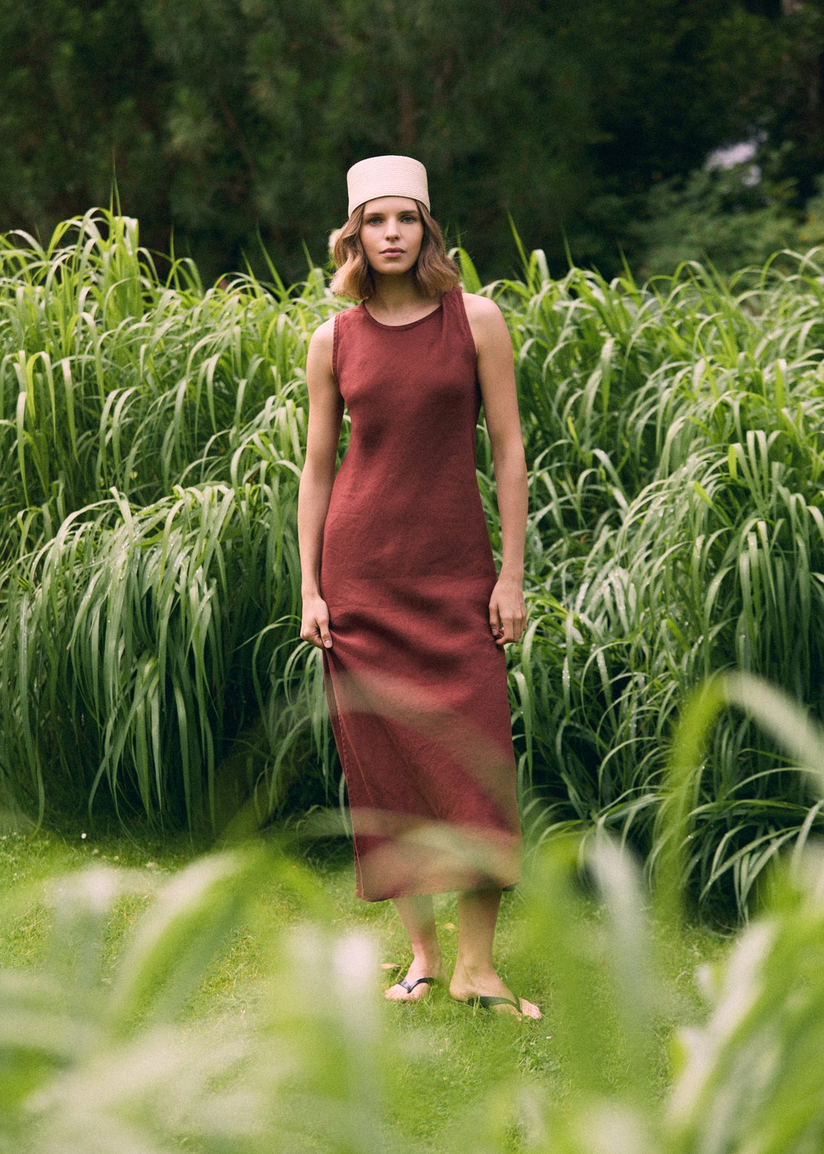 Woman wearing a terracotta sleeveless linen maxi dress and a light woven hat, standing among tall green grasses in a lush garden.