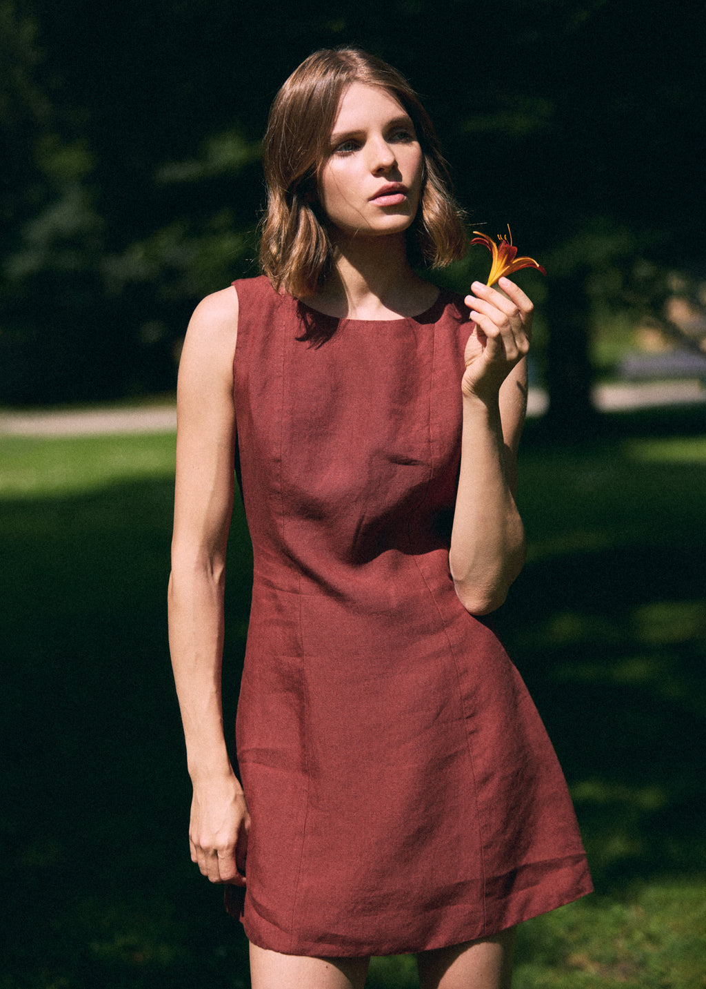 A woman wearing a sleeveless rust-red linen dress stands outdoors in a park, holding a small orange flower.