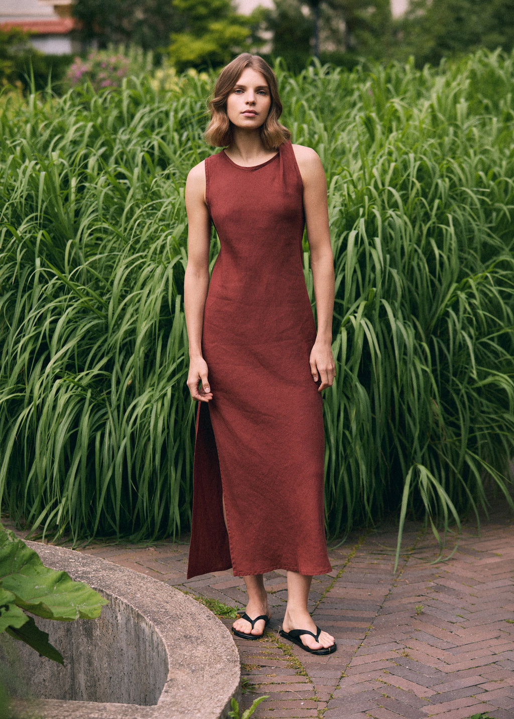 Woman wearing a terracotta sleeveless linen maxi dress with a subtle twisted shoulder detail and side slit, photographed against tall green grasses in a garden.