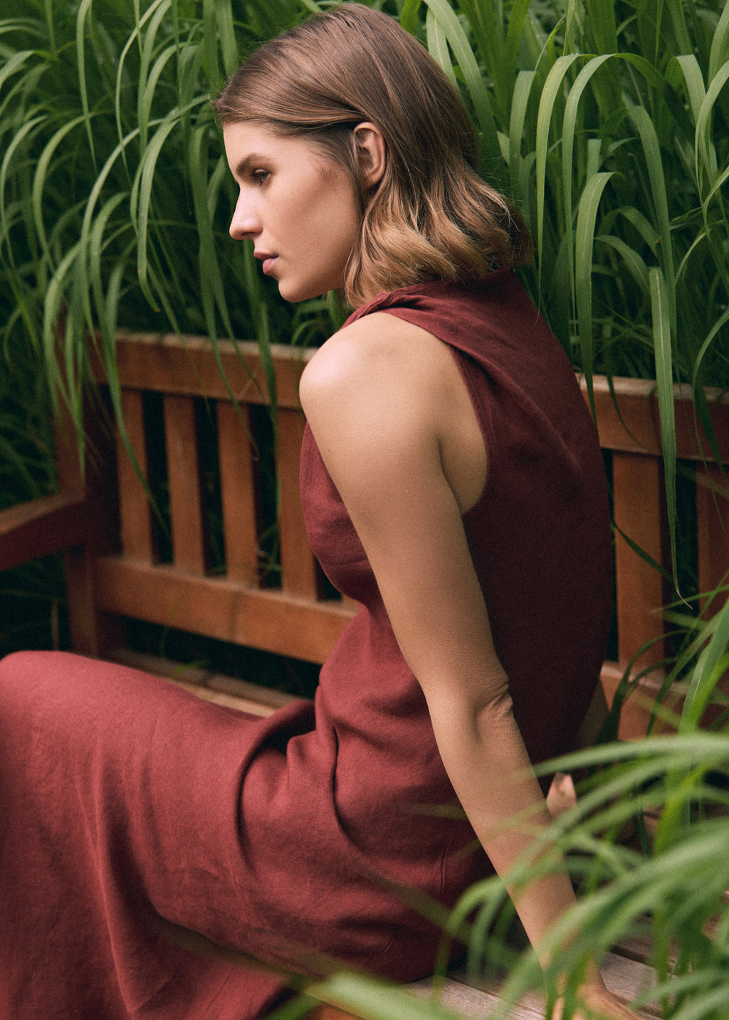 Close-up side view of a woman wearing a terracotta linen maxi dress while seated on a wooden bench, highlighting the natural linen texture, sleeveless cut and relaxed silhouette.