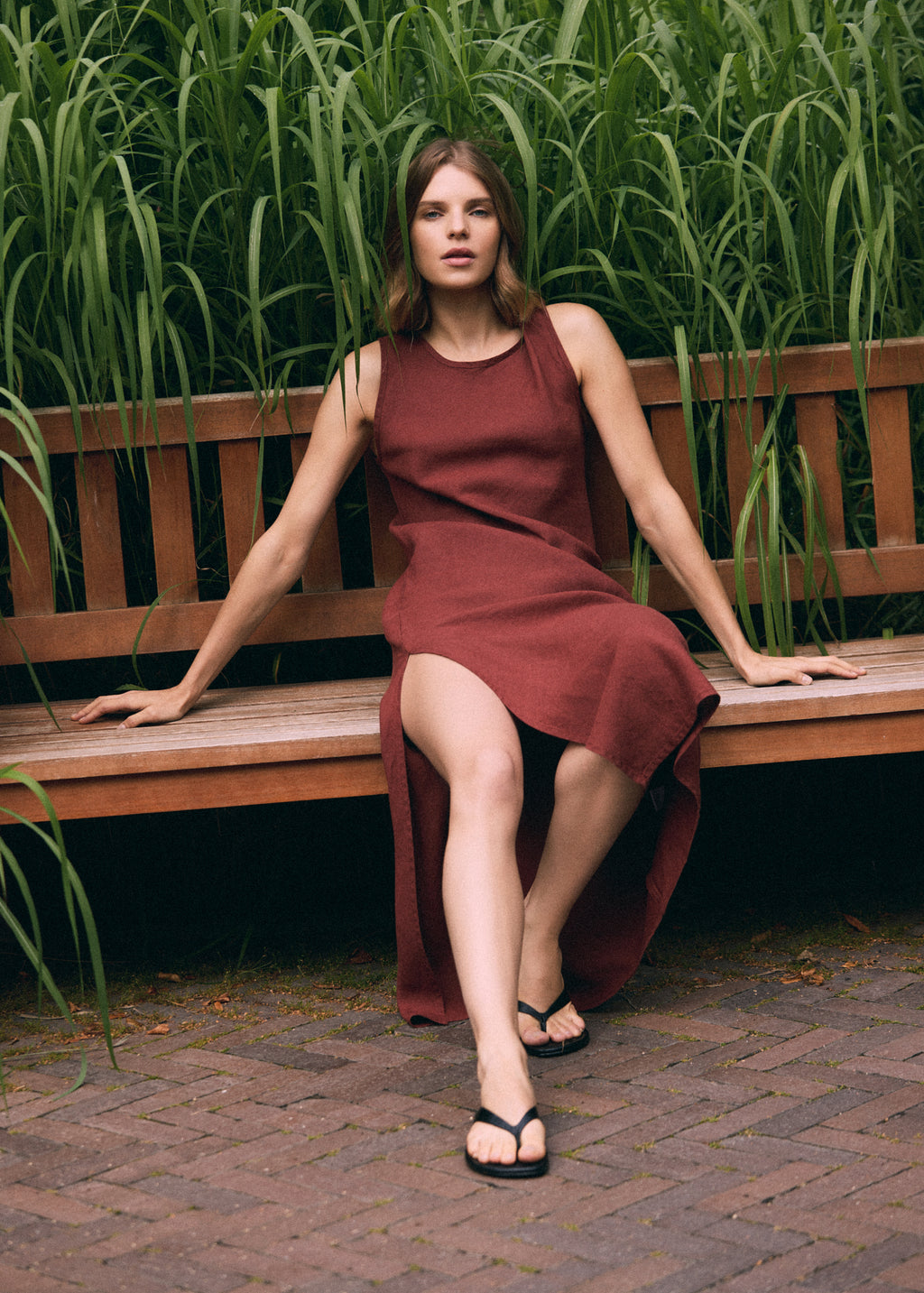 Woman in a terracotta linen maxi dress sitting on a wooden garden bench among lush tall grasses, showing the relaxed fit, flowing fabric and side slit of the dress.