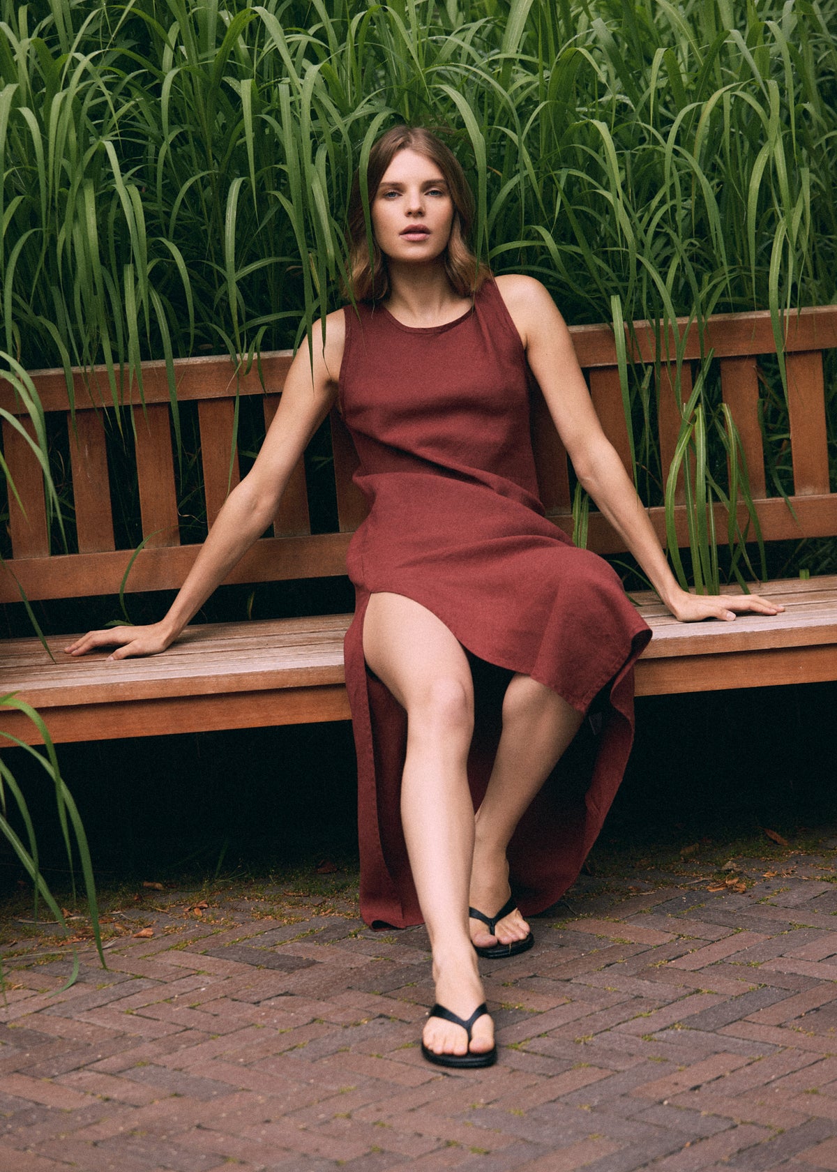 Woman in a terracotta linen maxi dress sitting on a wooden garden bench among lush tall grasses, showing the relaxed fit, flowing fabric and side slit of the dress.