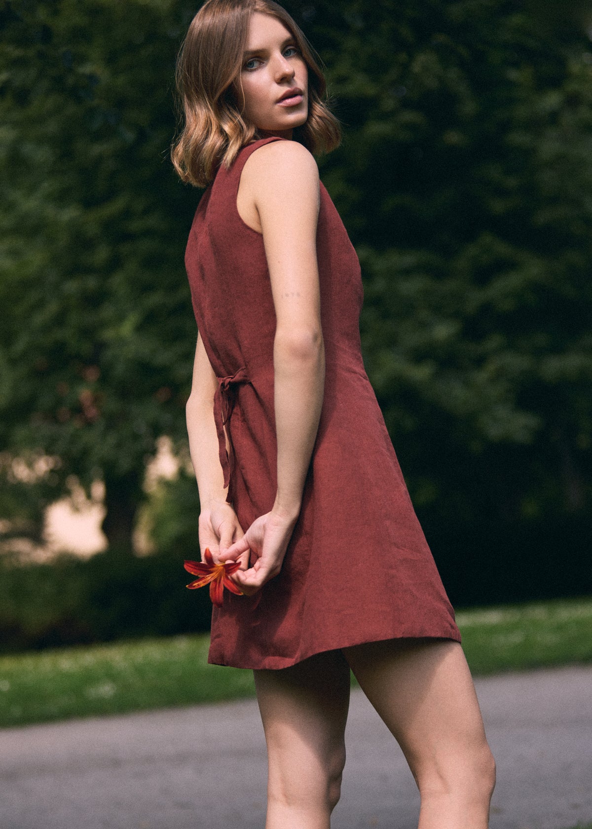 A woman wearing a sleeveless rust-brown linen dress, standing outdoors on a paved path with a blurred green garden background.