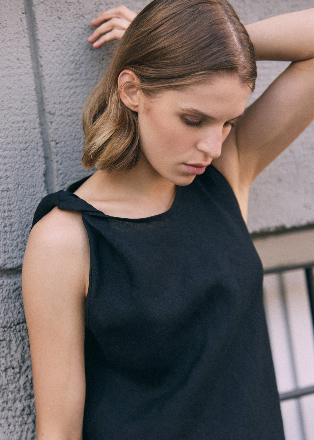 Close-up of a woman wearing a black sleeveless linen maxi dress with a distinctive twisted shoulder detail, photographed against a grey textured wall.