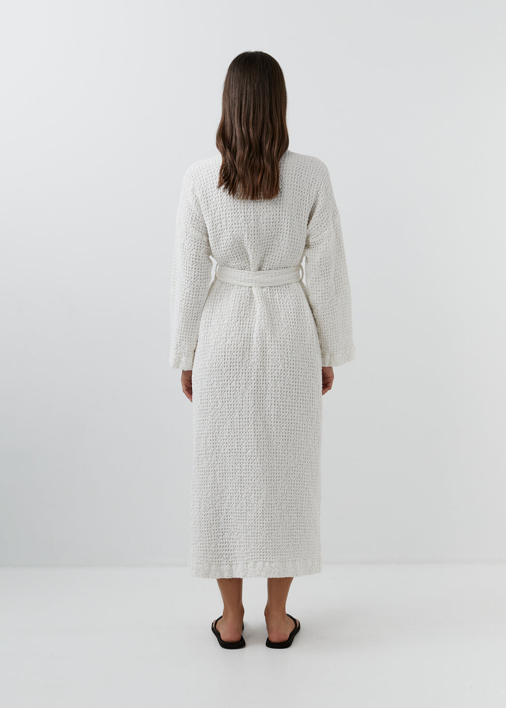 A woman viewed from behind wearing a white waffle-textured robe with a matching belt, standing against a plain white studio background.
