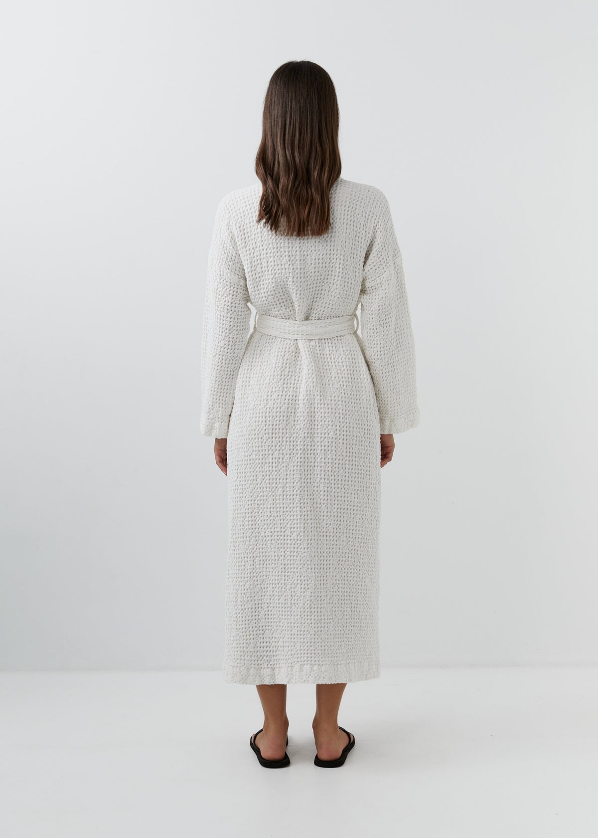 A woman viewed from behind wearing a white waffle-textured robe with a matching belt, standing against a plain white studio background.