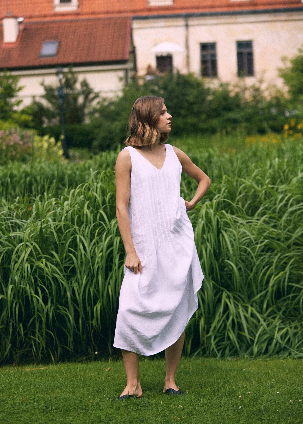 Woman standing in a green garden wearing a white sleeveless linen maxi dress with a V-neckline, pleated front and relaxed flowing silhouette, styled with black flats.