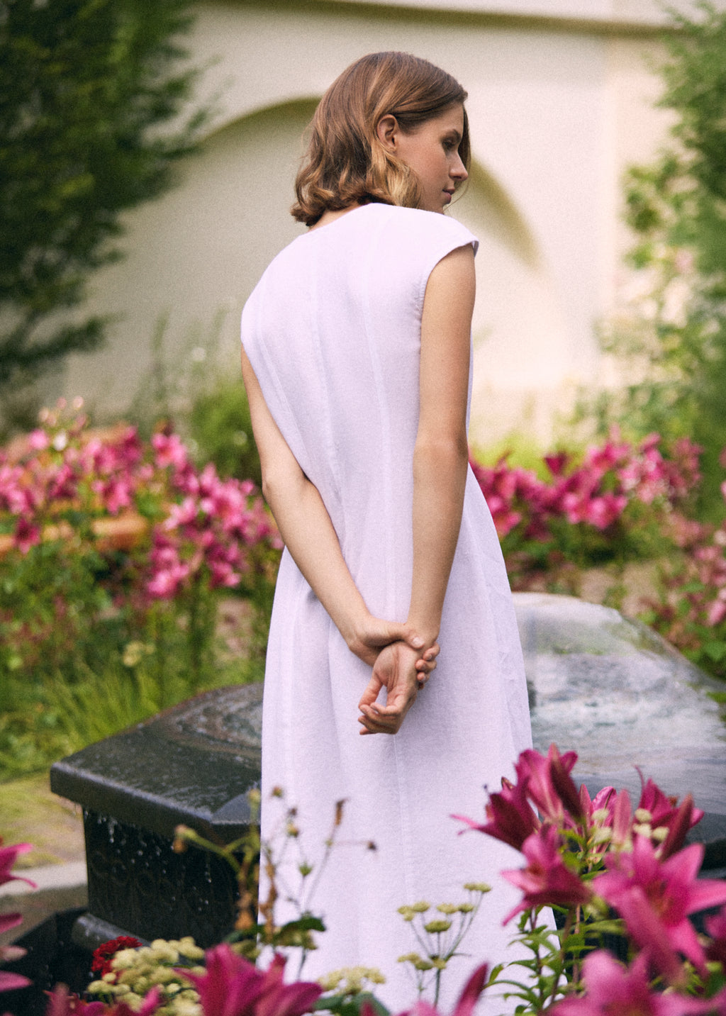 Woman wearing a white linen maxi dress with short cap sleeves and relaxed A-line silhouette, shown from the back in a blooming summer garden