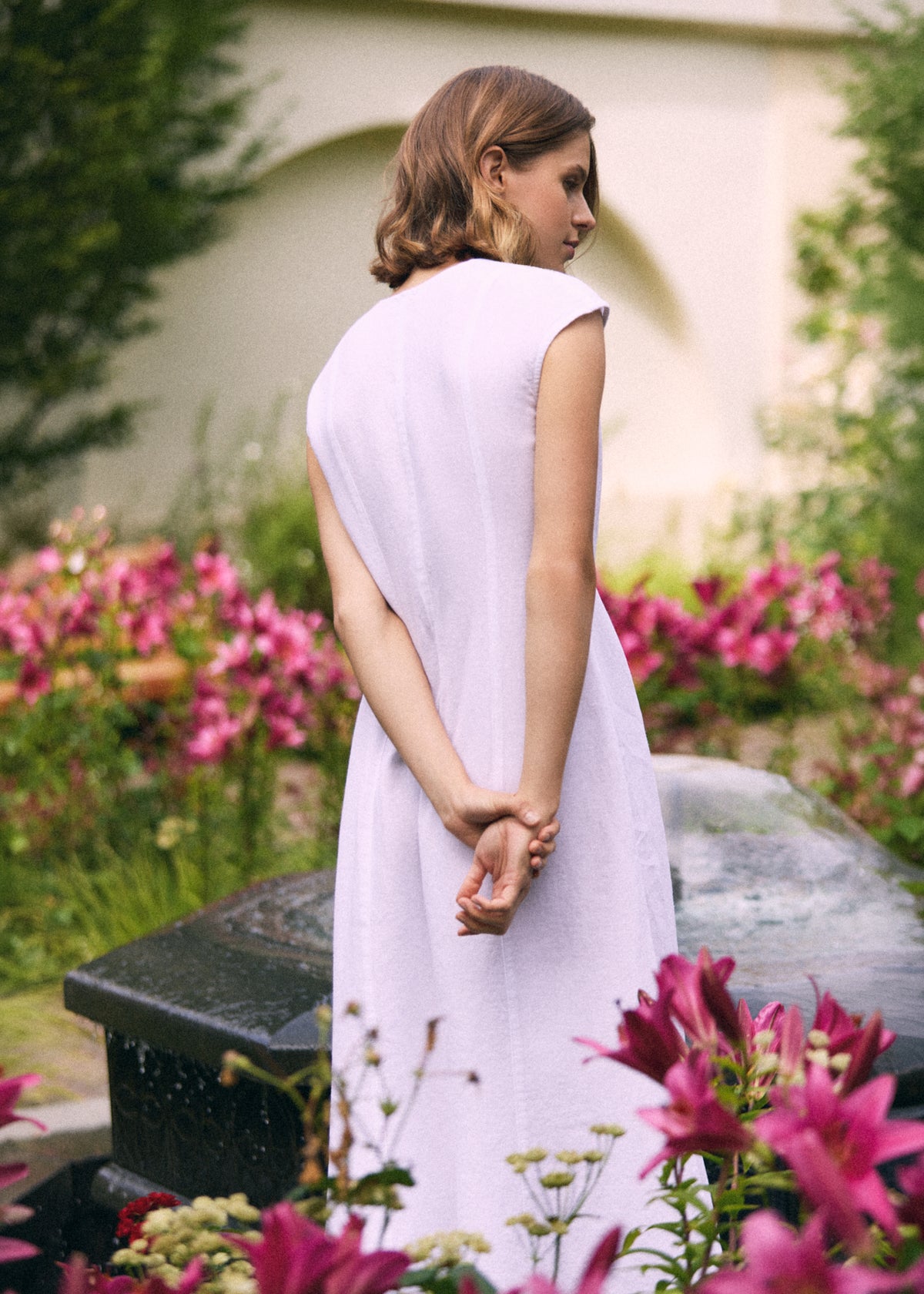 Woman wearing a white linen maxi dress with short cap sleeves and relaxed A-line silhouette, shown from the back in a blooming summer garden