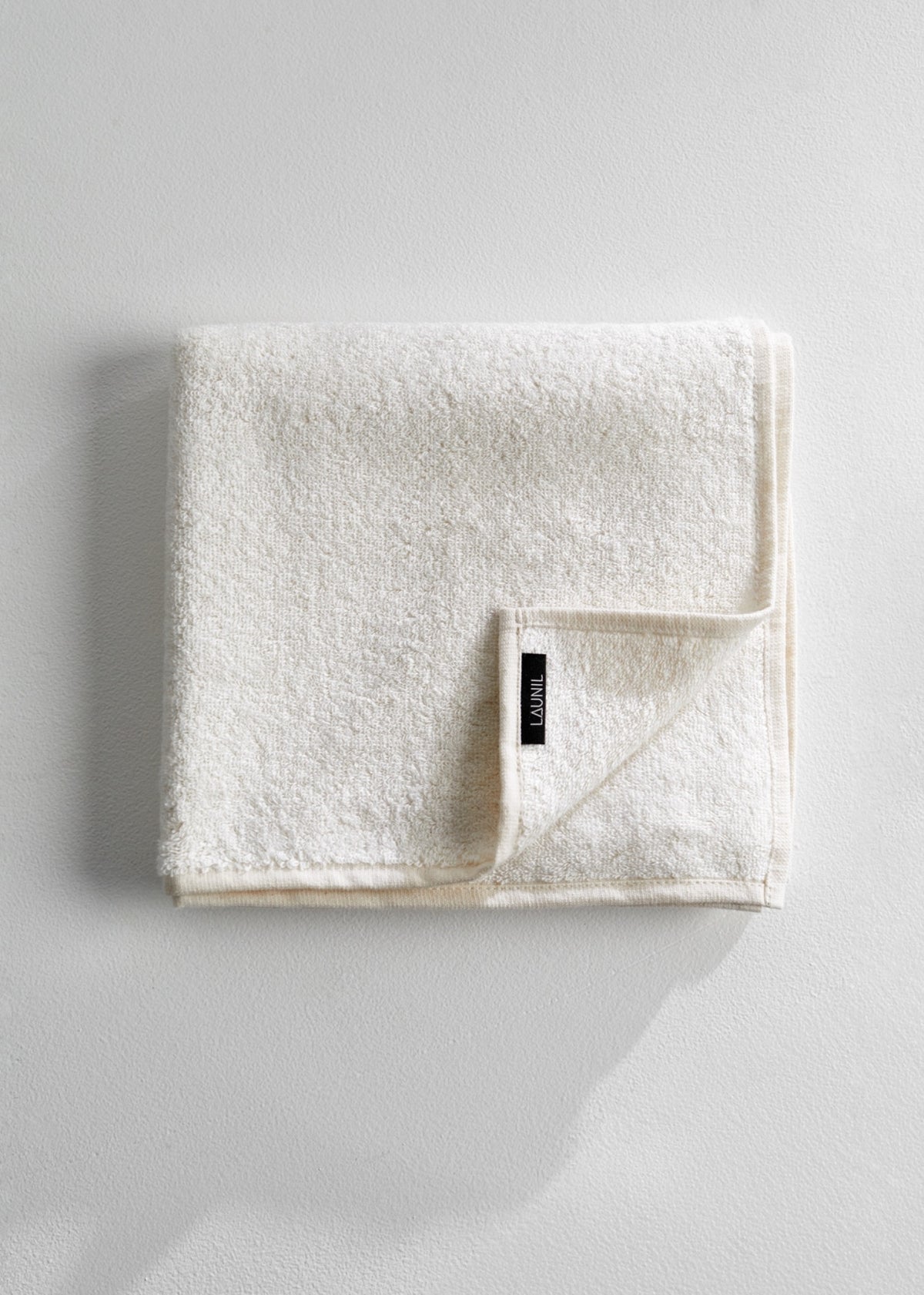 A folded off-white cotton towel with a small black brand tag, photographed on a light gray textured background.