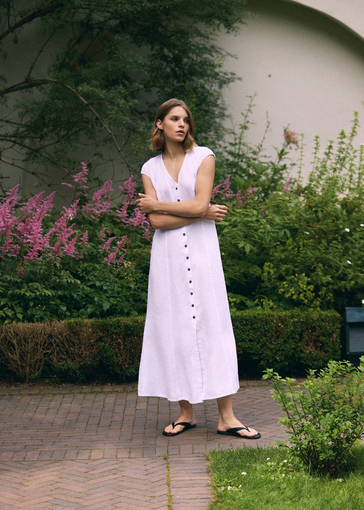 Woman wearing a white linen maxi dress with V-neck, front button closure, short cap sleeves and relaxed A-line silhouette, styled outdoors in a garden.