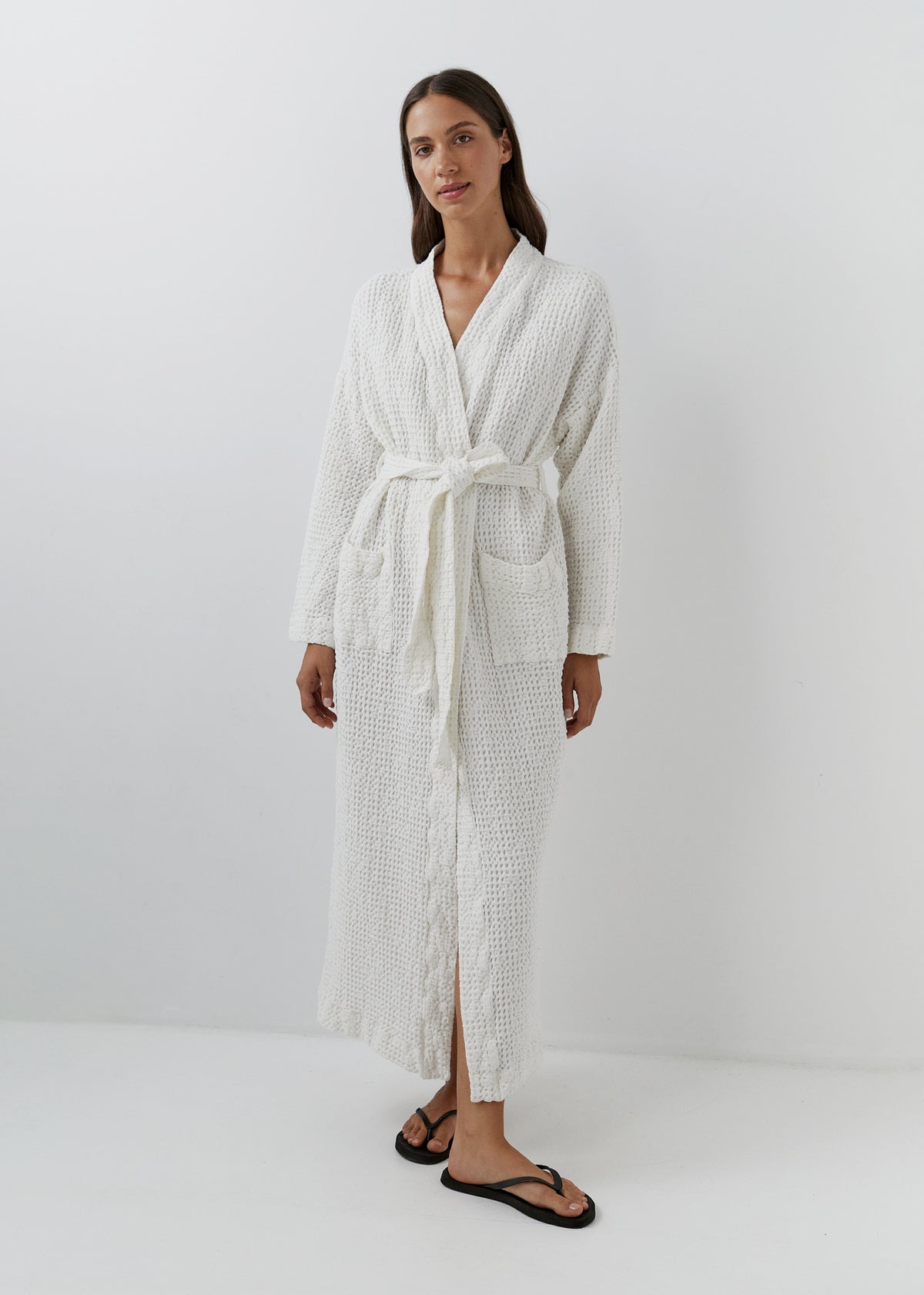 A woman modeling a long white textured bathrobe with a tie belt and front pockets, paired with black flip-flop sandals, against a plain white studio background.