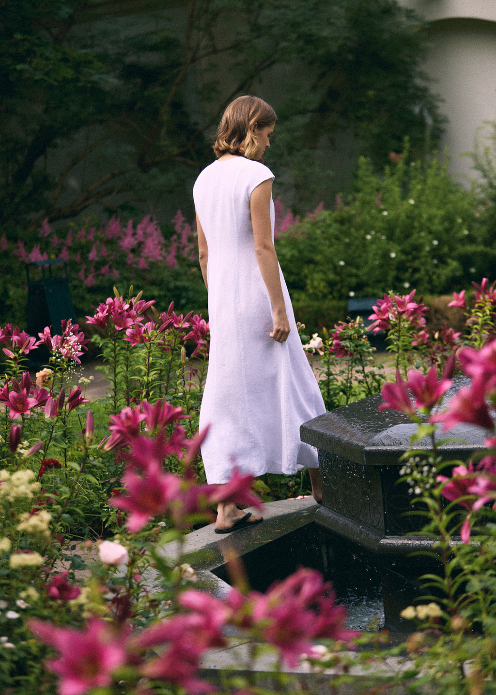 Woman wearing a white linen maxi dress with short cap sleeves and relaxed A-line silhouette, shown from the back in a blooming summer garden.