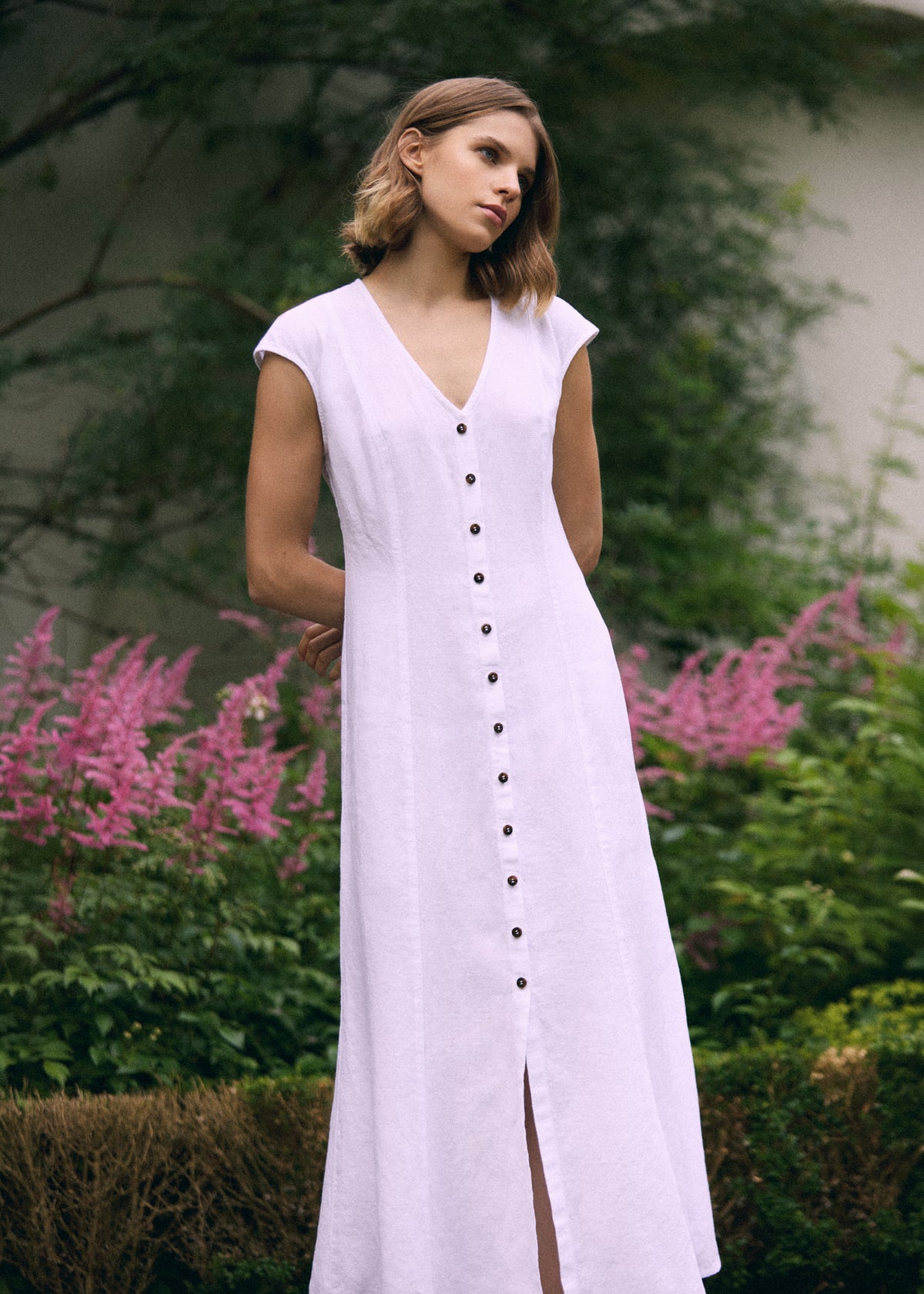 Woman wearing a white linen maxi dress with V-neck, dark front buttons, short cap sleeves and front slit, photographed outdoors in a summer garden.