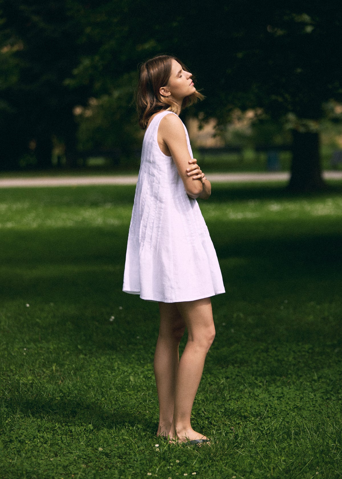 Woman standing on green grass in a white sleeveless linen mini dress with a loose A-line silhouette and above-the-knee length, shown from the side in a sunlit summer park.