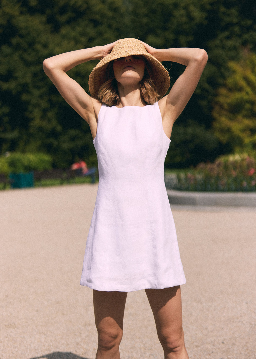 A woman wearing a sleeveless light lavender linen dress and a wide-brimmed straw hat, posing outdoors on a paved path with a blurred green garden background.