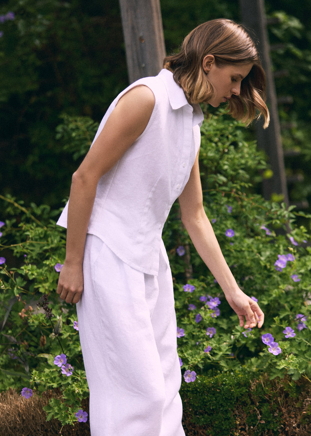 A woman wearing a white sleeveless collared top and matching wide-leg trousers stands outdoors in front of green foliage and purple flowers, looking down.