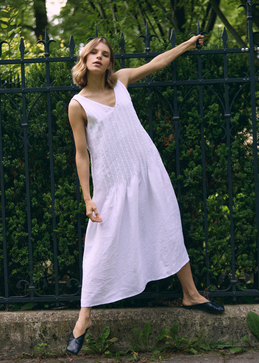 Woman wearing a white sleeveless linen maxi dress with a V-neckline, vertical pleated detailing and a flowing A-line skirt, posing by a black iron fence.