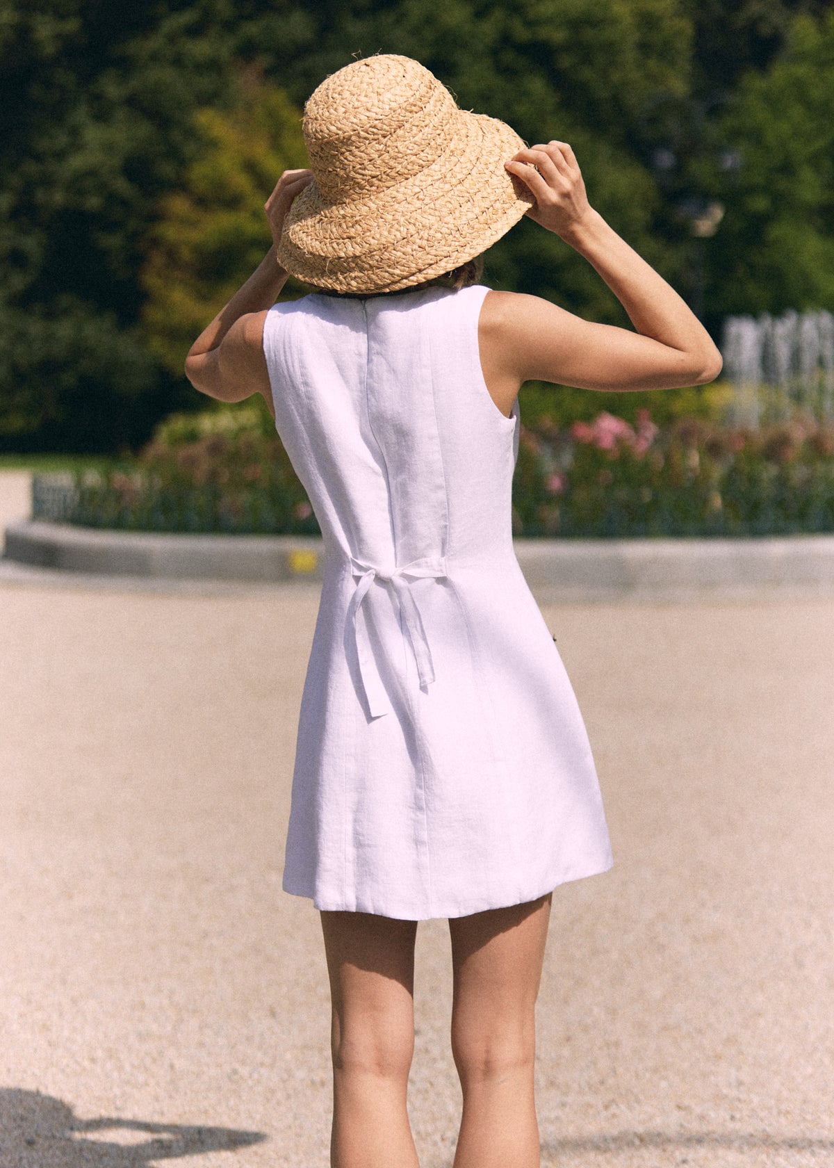 A woman viewed from behind wearing a sleeveless light lavender dress with a tie waist and a wide-brimmed straw sun hat, standing outdoors on a paved path with a blurred garden background.
