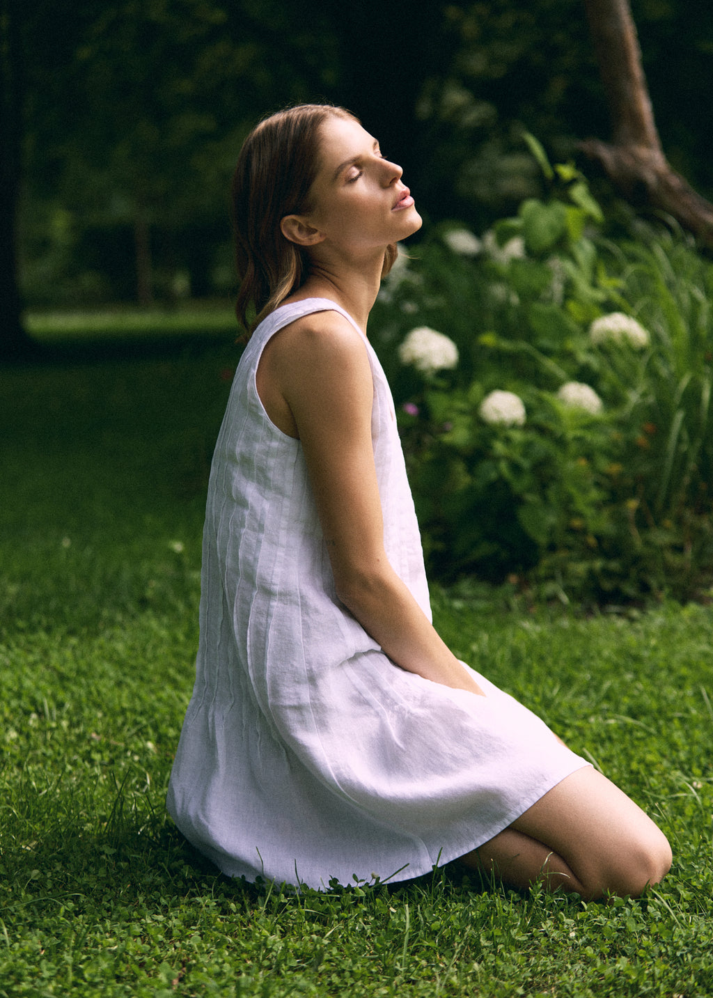 Woman sitting on green grass in a garden wearing a white sleeveless linen mini dress with a relaxed A-line silhouette and round neckline, shown from the side in a natural summer setting.